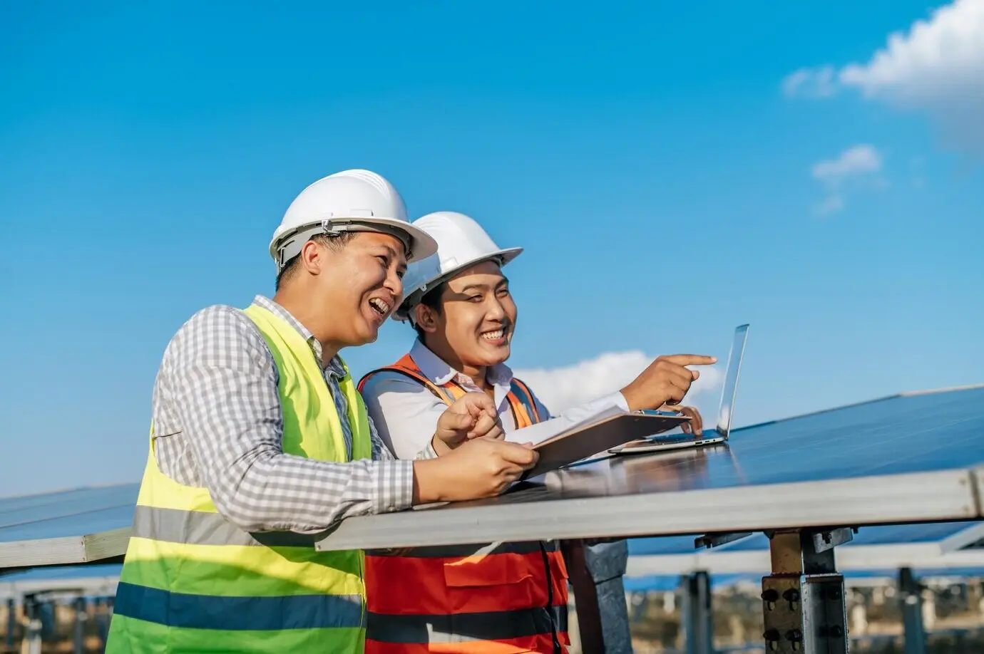 Two Asian men, a technician and an engineer, use a laptop while checking the sun’s efficiency as part of examining solar panel construction and talking together.