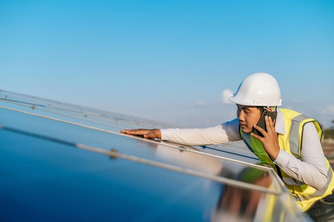 A young Asian male technician stands between long rows of photovoltaic solar panels, talking on a smartphone, with copy space.