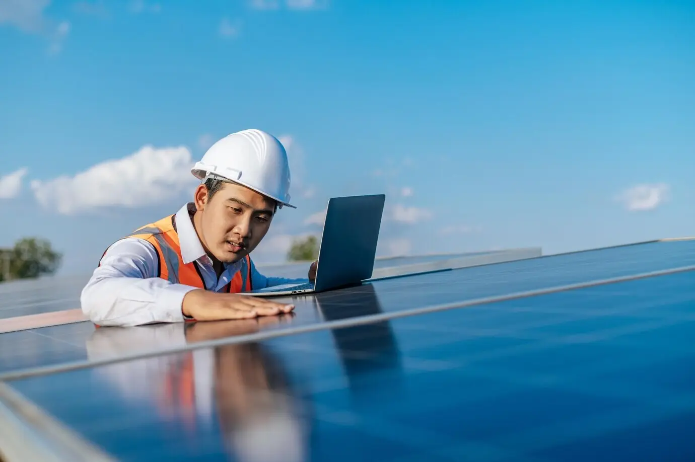 A young Asian male inspector-engineer and technician supervisor in a white helmet uses a laptop while working at a solar farm, checking the operation of the sun and photovoltaic solar panel at the station, with copy space.